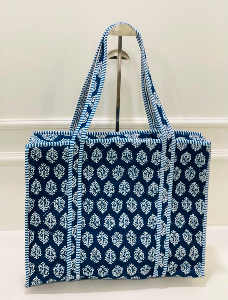 A blue and white patterned canvas tote bag with matching shoulder straps, displayed on a metal stand against a white wall.